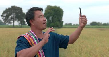 gelukkige aziatische boer man met lendendoek in een blauwe jurk video-oproep op smartphone en zwaaiende handen in het rijstveld. video