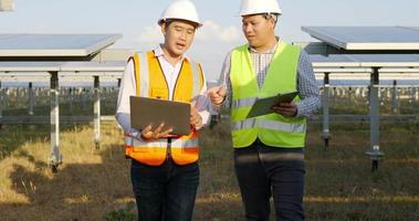 un ingeniero inspector asiático sostiene un tablero de control y un joven especialista usa una computadora portátil, dos ingenieros discuten y se alegran del éxito durante el trabajo en una granja solar, un panel solar en segundo plano