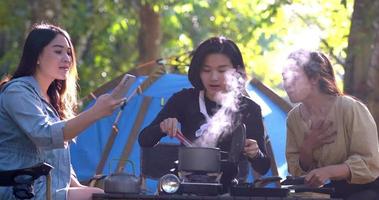 jonge aziatische vrouw die kookt en haar vriend geniet ervan om smartphone te gebruiken, maak een foto van de maaltijd in pot, ze praten en lachen samen met plezier tijdens het kamperen in het natuurpark