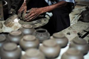 Selective focused on the dirty wrinkled skin hands of old man molding the clay work on the spinning wheel for making the jar with blurred group of clay jars in foreground photo