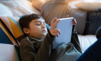 niño pequeño que se queda dormido mientras ve dibujos animados en una tableta, prueba a un niño acostado en la cama jugando en línea, niño durmiendo mientras lee un libro electrónico en una tableta digital, niño de la escuela relajándose en casa foto