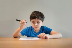 niño de la escuela usando un bolígrafo negro dibujando o escribiendo la carta en papel, un niño haciendo la tarea, un niño con un bolígrafo escribiendo notas en una hoja de papel durante la lección. alumno lindo haciendo la prueba, concepto de educación en el hogar foto