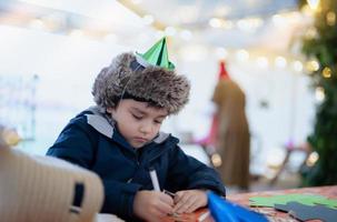 Cute boy with party hat using pink pen drawing or writing on paper colour with blurry bright light background, Kid having fun at birthday party, Child doing activity on Christmas or New Year holiday photo