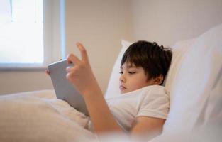 niño feliz acostado en la cama sosteniendo una tableta viendo dibujos animados y charlando con amigos en un bloc digital, un niño lindo jugando juegos en línea en Internet, un niño relajándose por la mañana antes de ir a la escuela foto