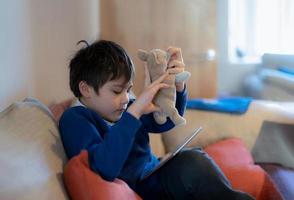 Happy schoolboy playing with dog toy and game online with friend on tablet,Kid using internet sending homework to the teacher, Positive child sitting on sofa relaxing  in living room in the morning photo