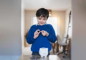 School kid putting coins into piggy bank, Child boy counting saving money, Young kid holding coin on his hands, Children learning financial responsibility and planning about saving money for future photo