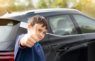 Happy young boy showing thumb up while stnading next to the car, School kid with smiling face standing at the carpark in sunny day Spring or Summer, Positive child going to school. photo