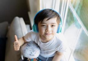 Happy young boy wearing headphones listening to music. Cute kid looking up to camera with smiling face, Positive Child sitting next to window relaxing on weekend in Spring or Summertime photo