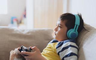 Portrait side view kid wearing headphones and playing video game. Child holding console play game online at home, Young boy siting sofa having fun and relaxing on his own on weekend photo