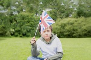 retrato de alta clave apuesto joven sosteniendo la bandera del reino unido, niño positivo con camiseta blanca sentado solo en el parque, un niño haciendo actividades al aire libre en primavera o vacaciones de verano foto