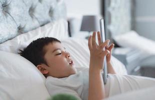 niño feliz acostado en la cama sosteniendo una tableta viendo dibujos animados y charlando con amigos en un bloc digital, un niño lindo jugando juegos en línea en Internet, un niño relajándose por la mañana antes de ir a la escuela foto