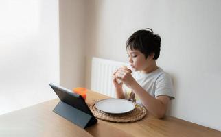 niño comiendo sándwiches de tocino caseros y viendo dibujos animados en una tableta, niño desayunando y jugando en línea en internet con amigos antes de ir a la escuela por la mañana foto