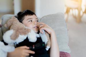 Happy Young boy holding remote control and looking up with smiling face, Cute kid lying on sofa enjoy watching TV program, Child relaxing at home on weekend photo