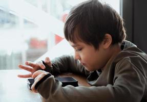 Hight key portrait kid playing game on mobile while waiting for food, Little boy sitting in coffee shop sending text to friends, Child playing game online on phone.Children with technology concept photo