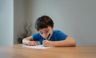 Kid siting on table doing homework,Child boy holding black pen writing on white paper,Young boy practicing English words at home. Elementary school and home schooling, Distance Education concept photo