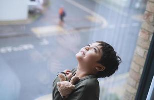Young kid standing next to window looking up to sky with smiling face, Child looking through window glass with wondering face. Portrait boy relaxing in room apartment in city photo