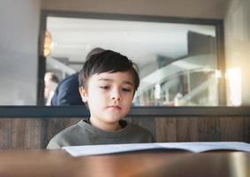 un niño de la escuela leyendo o eligiendo comida en el menú listo para pedir comida para el almuerzo, un niño sentado y esperando su comida en el restaurante. foto