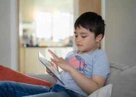 Kid sitting on sofa watching cartoon or playing game on tablet, Child boy using digital pad learning lesson online on internet, Home schooling,Distance learning online education concept photo