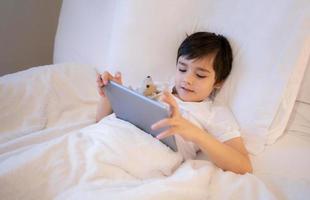 Happy Kid lying in bed holding tablet watching cartoon and chatting with friends on digital pad, Cute young boy playing games online on internet, Child relaxing in the morning before go to school photo