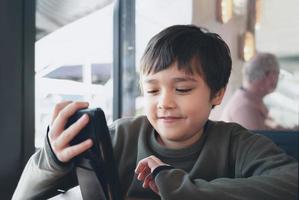 Cenimatic portrait young boy playing game on mobile while waiting for food, Kid sitting in coffee shop sending text to friends, Child playing game online on phone. photo
