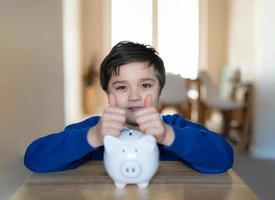 Happy School boy putting thumbs up on piggy bank and looking at camera with smiling face.Child showing money saving box.kid Learning financial responsibility and planning about saving money for future photo