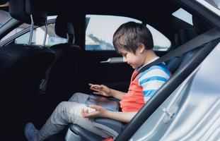 retrato cinematográfico niño feliz usando una tableta mientras está sentado en el asiento trasero del pasajero con un cinturón de seguridad, niño escribiendo en una almohadilla inteligente, niño de la escuela que viaja a la escuela en automóvil. regreso a la escuela foto