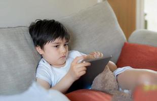 Young boy holding tablet looking out deep in thought, Unhappy kid with sad face sitting alone on sofa, A lonely child playing with soft toy and playing game on digital pad at home. photo