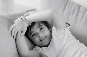 Portrait black and white image of happy young boy looking at camera with smiling face, Positive child lying on sofa relaxing at home. Head shot mono tone of smile kid photo