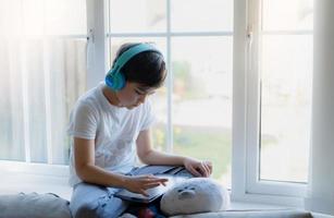Happy young boy wearing headphone for playing game online on internet with friends, Kid sitting next to window reading or watching cartoon on tablet Child relaxing at home in the morning on Springtime photo