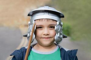 Happy boy wearing lobster-tailed pot helmet costume pretending to be a legionary  soldier, Active kid playing outside in the park field on Spring, Child having fun doing outdoors activity in  Summer photo