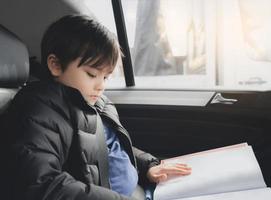 Kid siting on car seat and reading a book, Young boy sitting in  safety seat, Portrait of child entertaining himserf on a road trip. Concept of safety taveling by car with children photo