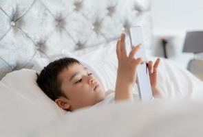 niño feliz acostado en la cama sosteniendo una tableta viendo dibujos animados y charlando con amigos en un bloc digital, un niño lindo jugando juegos en línea en Internet, un niño relajándose por la mañana antes de ir a la escuela foto