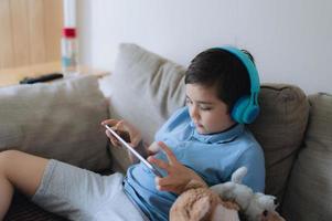 niño feliz usando auriculares para jugar en Internet con amigos, niño sentado en el sofá leyendo o viendo dibujos animados en la tableta, niño de la escuela haciendo la tarea en línea en casa. foto
