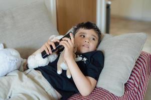 Happy Young boy holding remote control and looking up with smiling face, Cute kid lying on sofa enjoy watching TV program, Child relaxing at home on weekend photo
