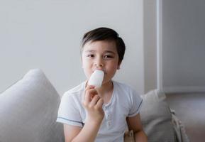 niño feliz comiendo paleta de hielo, retrato de un niño lindo sentado en un sofá al lado de una ventana tomando un refresco, niño con cara sonriente relajándose en casa. niño mirando a la cámara mientras come helado foto