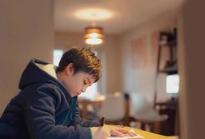 niño sentado en la mesa haciendo la tarea, niño sosteniendo un bolígrafo negro escribiendo en papel blanco, niño practicando palabras en inglés en casa. escuela primaria y educación en el hogar, concepto de educación a distancia foto