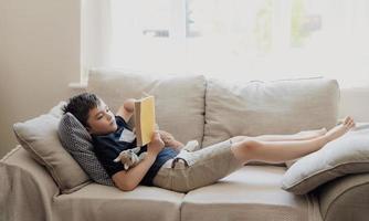 niño de la escuela leyendo un libro para la tarea, niño de larga duración acostado en el sofá disfruta leyendo la historia en la sala de estar, niño relajándose en casa en el verano soleado, educación en el hogar, concepto de educación foto