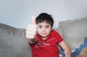 niño joven alegre y feliz con camiseta roja que muestra el pulgar hacia arriba, niño de la escuela que muestra el signo de ok y mira la cámara, enfoque selectivo en la cara. foto