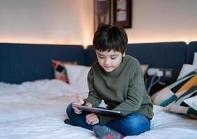 niño viendo dibujos animados y charlando con amigos en una tableta, niño feliz sentado en la cama jugando juegos en línea en una libreta digital, niño retratado relajándose en casa en su dormitorio el fin de semana. foto