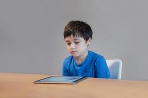 niño de la escuela usando una tableta haciendo la tarea en línea, niño con la cara aburrida leyendo un libro en un lector digital, niño leyendo una historia en un libro electrónico, educación en el hogar, aprendiendo el concepto de educación en línea foto