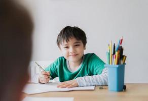 Happy boy using pencil drawing or sketching on paper, Cute kid with smiling face siting on table doing homework, Child enjoy art and craft activity with friends, Education concept photo