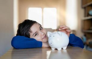 niño de la escuela poniendo monedas en una alcancía, niño contando dinero ahorrado, niño pequeño sosteniendo una moneda en sus manos, niños aprendiendo responsabilidad financiera y planeando ahorrar dinero para el futuro foto