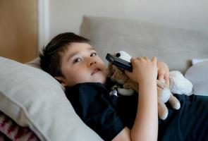 Portrait young boy holding remote control and looking up with curious face, Kid sitting on sofa watching cartoon on TV, Child lying down on couch relaxing in living room after back from school. photo