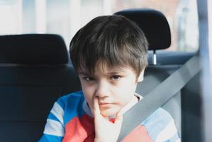 retrato de niño con cara aburrida, cara triste de niño sentado en el asiento trasero del pasajero con un cinturón de seguridad, niño infeliz mirando hacia afuera, concepto de niños mimados foto