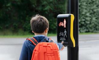 niño de la escuela de retrato de vista trasera presionando el botón en los semáforos en el cruce de peatones de camino a la escuela. niño con mochila usando instalaciones peatonales controladas por señales de tráfico para cruzar la calle foto