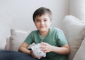 Happy boy holding piggy bank with smiling face. Indoor portrait of a cheerful child showing money saving box.School kid Learning financial responsibility and planning about saving for future concept photo