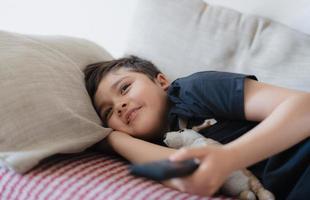 Happy Young boy holding remote control and looking up with smiling face, Cute kid lying on sofa enjoy watching TV program, Child relaxing at home on weekend photo