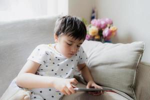 High key portrait kid playing game on tablet sitting on sofa with light shining from window, Young boy  playing games on internet, Cinematic portrait Child doing homework online at home photo