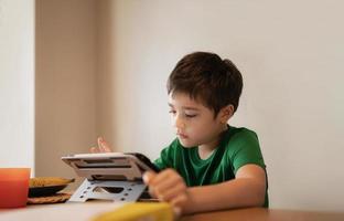 niño jugando en línea con amigos en tableta, retrato cinematográfico joven videollamada con amigos mientras desayuna, niño viendo dibujos animados en Internet, niño de la escuela haciendo la tarea en la libreta digital foto