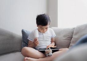 Kid eating coconut ice lolly while playing game on tablet,Hight key portrait Child sitting on sofa doing homework online at home,A boy playing games on internet with light shining from window photo